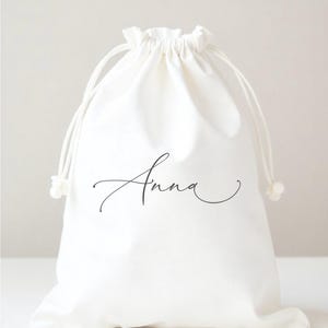May include: A white fabric drawstring pouch featuring the name Anna printed in a black cursive script font. The bag has a gathered top closure with two white cords that pull to secure the opening, creating a simple and functional storage container.