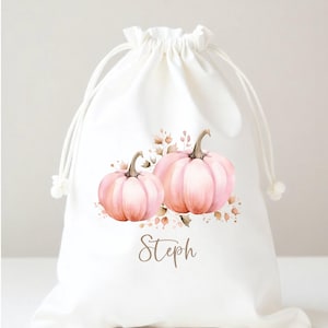 May include: A white drawstring bag featuring a watercolor design of two pink pumpkins with brown stems and leaves. The name "Steph" is written in a cursive font below the pumpkins. The bag is tied with white cords.