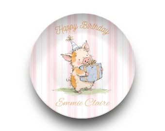 May include: A round ceramic plate featuring a light pink and white vertical striped background. The center displays an illustration of a piglet wearing a blue and white party hat, holding a blue gift box. Text reads Happy Birthday and Emmie Claire.