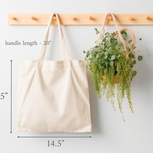 May include: A cream-colored tote bag with a 20-inch handle length, 15-inch height, and 14.5-inch width hangs next to a hanging basket filled with green plants. The tote bag is made of a plain, unadorned fabric.