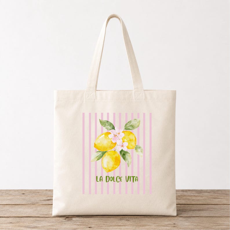 May include: Cream-colored tote bag featuring a watercolor illustration of lemons and blossoms. The design includes pink vertical stripes and the text "LA DOLCE VITA" in green. The bag has a simple, rectangular shape with two handles.