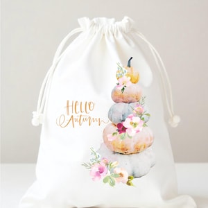 May include: White drawstring bag featuring a watercolor design of stacked pumpkins in shades of orange, gray, and yellow, adorned with pink flowers and green leaves. The words "Hello Autumn" are written in gold script.