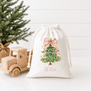Puede incluir: Una bolsa blanca con cord&oacute;n que presenta una ilustraci&oacute;n impresa de un &aacute;rbol de Navidad verde adornado con un gran lazo rosa y luces. El nombre Stella est&aacute; impreso en letra cursiva rosa debajo del dise&ntilde;o del &aacute;rbol en la parte frontal.