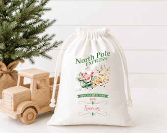 May include: A white drawstring gift bag featuring a watercolor illustration of Santa Claus in a sleigh pulled by reindeer. The text reads North Pole Express, Special Delivery For, and the name Seamus in red script with green decorative accents.