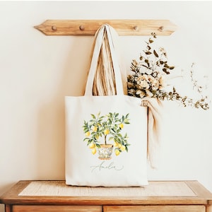 May include: A cream tote bag with a watercolour illustration of a lemon tree in a decorative pot. The tree is full of yellow lemons and green leaves. The name "Amelia" is written in script below the tree. The bag hangs on a wooden rack.