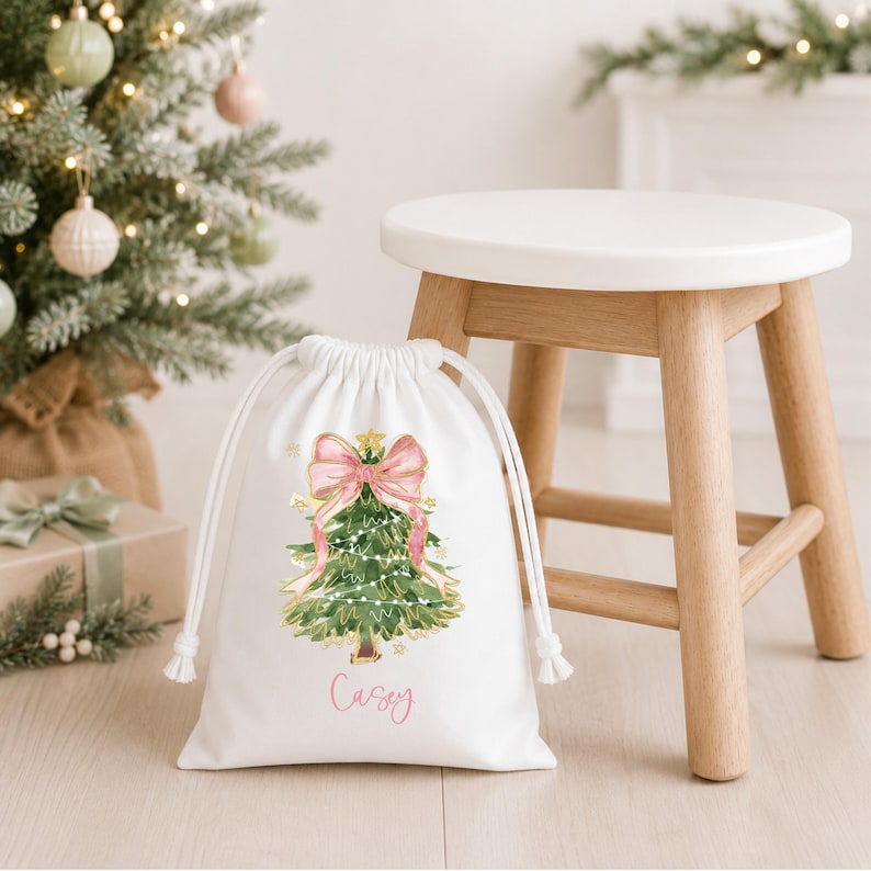 Puede incluir: Una bolsa de regalo blanca con cord&oacute;n que presenta una ilustraci&oacute;n en acuarela de un &aacute;rbol de Navidad verde decorado con un gran lazo rosa y luces. El nombre Casey est&aacute; impreso en rosa debajo del &aacute;rbol. La bolsa tiene cordones blancos.