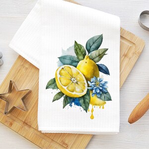 K&ouml;nnte beinhalten: Ein wei&szlig;es K&uuml;chentuch mit einer Aquarell-Illustration von Zitronen und einer blauen Blume. Die Zitronen sind gelb und haben gr&uuml;ne Bl&auml;tter. Die Blume ist blau mit einer gelben Mitte. Das Tuch liegt auf einem Holzbrett mit einem Nudelholz und Ausstechern.