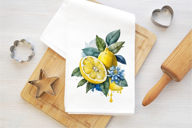 K&ouml;nnte beinhalten: Ein wei&szlig;es K&uuml;chentuch mit einer Aquarell-Illustration von Zitronen und einer blauen Blume. Die Zitronen sind gelb und haben gr&uuml;ne Bl&auml;tter. Die Blume ist blau mit einer gelben Mitte. Das Tuch liegt auf einem Holzbrett mit einem Nudelholz und Ausstechern.
