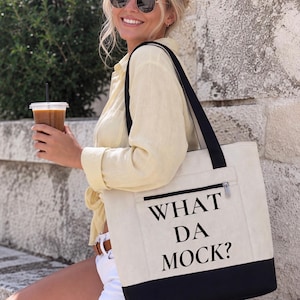 Peut inclure: Un sac fourre-tout en toile couleur crème avec une base noire et des bretelles noires. Le sac présente une poche frontale zippée et le texte imprimé en noir WHAT DA MOCK? centré sur le panneau avant dans une police serif en gras.