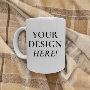 May include: A white ceramic mug with a curved handle. The mug features the text "YOUR DESIGN HERE!" in a bold, black font. The mug is set against a tan and brown plaid blanket.