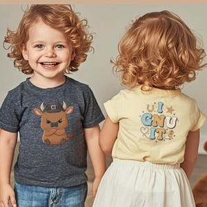 Puede incluir: Dos niños con camisetas. Uno lleva una camiseta gris oscuro con un dibujo de un toro de dibujos animados y vaqueros azules. El otro lleva una camiseta amarilla con el texto "U I GNU IT" en la espalda y una falda blanca.