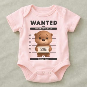 Könnte beinhalten: Hellrosa Baby-Body mit dem Text "WANTED FOR CHEEKY CUDDLES". Ein Cartoon-Bär hält ein Schild mit dem Namen "Sofia". Das Design ist für ein Baby.