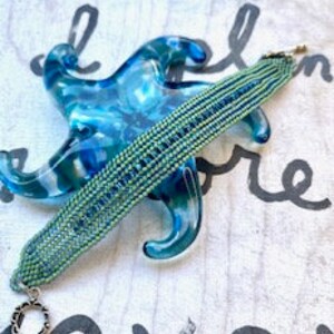 May include: A green and blue beaded bracelet with silver clasps. The bracelet is woven with small beads in a geometric pattern.