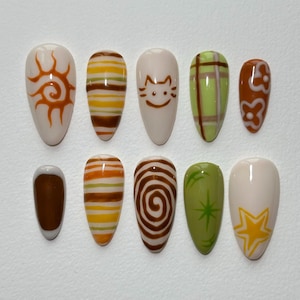 May include: A set of ten almond-shaped press-on nails with various designs. The designs include a sun, stripes, a cat face, plaid, flowers, a swirl, a star, and a green starburst. The nails are painted in shades of brown, yellow, green, and white.