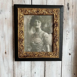 Peut inclure: Portrait encadré noir et or d'une femme en robe d'époque. Le portrait est en noir et blanc, la femme portant un collier et une coiffe décorative. Le cadre présente un motif doré orné.