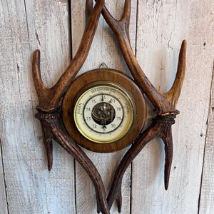 Op de afbeelding: Een vintage barometer met een houten frame en een crèmekleurige wijzerplaat, gemonteerd op decoratieve geweien. De wijzerplaat heeft een naald en het woord "VERANDERLICH". De geweien hebben een diepbruine kleur.
