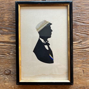 Peut inclure: Silhouette encadrée représentant un profil noir d'une personne en costume et casquette. L'œuvre d'art a un cadre doré et noir, et le fond est de couleur beige clair. La casquette a un design beige et doré.