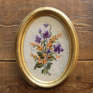 Könnte beinhalten: Eine gerahmte Stickerei mit einem Strauß aus lila und orangefarbenen Blumen auf weißem Hintergrund. Der Rahmen ist oval und golden.