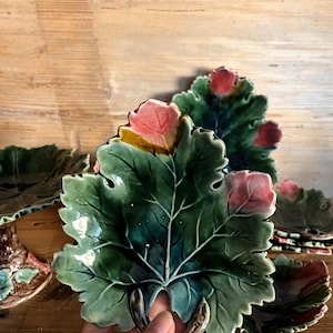 Op de afbeelding: Een keramieken bord in de vorm van een groen druivenblad met opvallende nerven en roze bloemknoppen. De glanzende afwerking benadrukt het textuurrijke oppervlak van het blad. Het bord heeft een kleine, steeltje-achtige handgreep aan de basis.