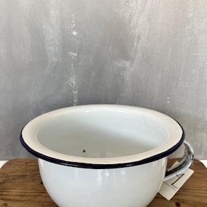 Op de afbeelding: Witte emaille kom met een donkerblauwe rand en handvat. De kom staat op een houten oppervlak tegen een grijze achtergrond. Een klein label is aan het handvat bevestigd.