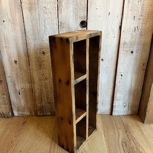 Könnte beinhalten: Eine rechteckige Holz-Aufbewahrungsbox mit drei Innenregalen. Die Box besteht aus hellbraunem Holz mit sichtbarer Maserung und Knoten und hat ein verwittertes Aussehen. Die Box ist für die Aufbewahrung kleiner Gegenstände konzipiert.