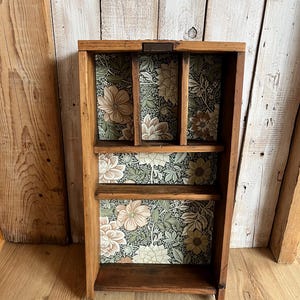 May include: A wooden shelf unit with three shelves and a floral patterned backing. The unit is made of dark brown wood and has a rectangular shape. The floral pattern includes beige and green flowers.