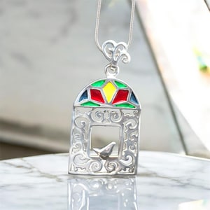 May include: A silver-colored pendant necklace featuring a bird in a cage design. The cage has colorful stained-glass-style accents in red, yellow, green, and blue. The pendant hangs from a delicate silver chain.