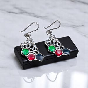 May include: A pair of silver-toned earrings with a filigree design. Each earring features three colorful, hexagonal accents in green, red, and navy blue. The earrings are displayed on a black surface.