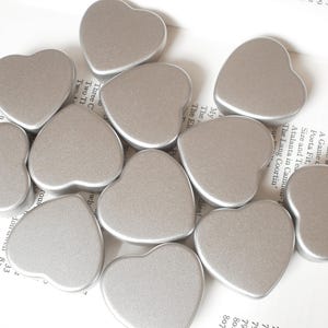 Peut inclure: Une collection de boîtes en forme de cœur argentées. Les boîtes sont disposées en grappe sur une surface blanche.
