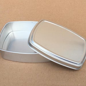 Può includere: Un contenitore rettangolare in metallo argentato con un coperchio. Il contenitore è aperto e il coperchio è posto accanto.