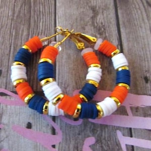 May include: A pair of hoop earrings made with colorful polymer clay beads. The beads are orange, white, and blue, and are arranged in a repeating pattern. The earrings are gold-toned and have a clip-on closure.