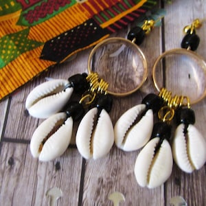 May include: A pair of gold hoop earrings with black beads and white cowrie shells hanging from them. The earrings are on a wooden surface.