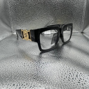 Puede incluir: Gafas de montura cuadrada negra con lentes transparentes y bordes gruesos. Las patillas están decoradas con un prominente emblema circular en tono dorado que muestra una figura en relieve detallada, uniendo el frente con las patillas.