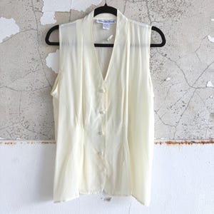 May include: A sleeveless cream-colored vest featuring a V-neckline, a four-button front closure with matching cream buttons, and vertical seam detailing. The garment is displayed on a black hanger and includes a label reading Ann and Frank.