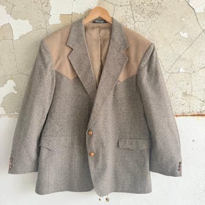 Puede incluir: Blazer gris con detalles de hombros en ante beige y botones de madera. La chaqueta está colgada de una percha de madera. El blazer tiene dos bolsillos delanteros y botones en las mangas. Fondo de pared desgastada.