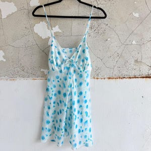 May include: A white sleeveless mini dress featuring a repeating pattern of small blue flowers. The garment has thin spaghetti straps, a V-neckline with a small central bow detail, and a lightweight, flowing silhouette that hangs from a black hanger.