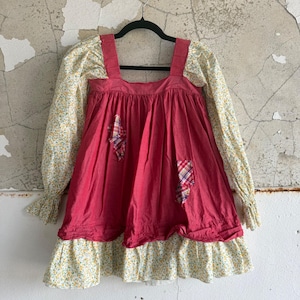 Puede incluir: Un vestido de manga larga con un cuerpo de color rosa oscuro liso, escote cuadrado y dos pequeños parches de tela a cuadros. Las mangas y el volante inferior están hechos de tela amarilla clara con un pequeño estampado floral azul y verde.