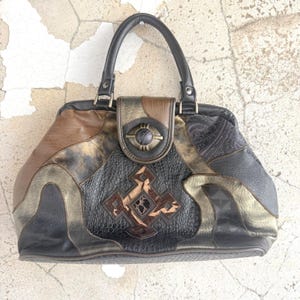 May include: A structured handbag featuring a patchwork design of black, brown, and metallic gold panels. The front displays a central geometric emblem with a dark circular clasp above. It includes a single black handle and varied textures.