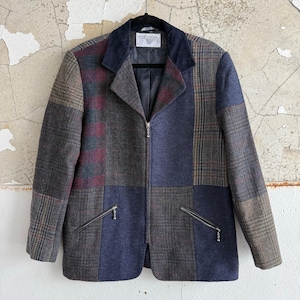 Peut inclure: Veste patchwork avec un col bleu marine et une fermeture éclair intégrale. La veste présente un mélange de tissus à carreaux et unis dans des tons de bleu, marron et bordeaux. Deux poches zippées sont visibles sur le devant. La veste est suspendue à un cintre noir.