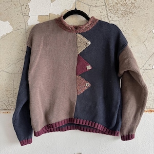 Puede incluir: Un suéter de bloques de color con un diseño único. El suéter presenta una combinación de tonos marrón, azul marino y burdeos. La parte delantera tiene detalles geométricos con botones plateados. El cuello y los puños son acanalados, y el suéter está colgado de una percha negra.