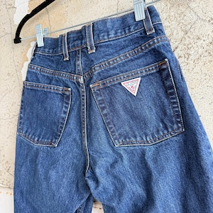 May include: Close-up of the rear of blue denim jeans. The jeans have two back pockets and a small red and white triangle logo on the right pocket. The jeans are hanging on a black hanger.