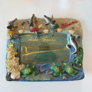 May include: A decorative resin picture frame with a Hawaiian theme. The frame features dolphins, turtles, and the text "Aloha Hawaii." The frame has a sandy texture and a blue water design.