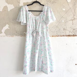 May include: A light blue dress featuring a repeating floral pattern in pink and green. The garment has short flutter sleeves, a scoop neckline trimmed with white lace, and an elasticized waist with a matching fabric tie centered at the front.