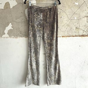 May include: A pair of high-waisted trousers featuring a repeating grid pattern in shades of brown, tan, and black. The trousers have a fitted waist and a flared leg silhouette, hanging from a black metal hanger against a textured wall surface.