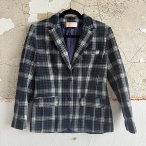 May include: A plaid blazer in shades of navy, green, white, and purple hangs on a black hanger. The blazer has a two-button closure, a notched lapel, and two front pockets. The interior lining is a solid navy blue.