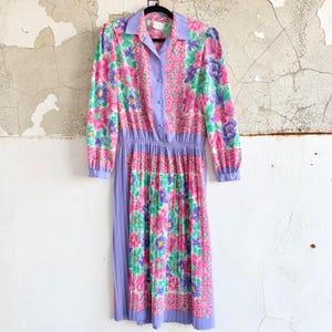 May include: A vintage floral print dress with long sleeves and a pleated skirt. The dress features a collared neckline and a button-down bodice in a lavender color. The floral pattern includes pink, purple, and green flowers on a white background.