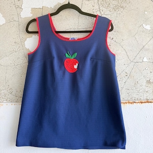 Könnte beinhalten: Ein ärmelloses, geripptes Tanktop in Marineblau mit einem Rundhalsausschnitt, der mit rotem Keder verziert ist. In der Mitte der Brust befindet sich eine aufgestickte rote Apfel-Applikation mit grünen Blättern und einem kleinen weißen Biss.