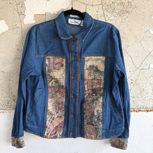 May include: A blue denim jacket featuring a button-down front with brown piping and two vertical panels of beige fabric printed with a multi-colored floral and garden scene. The jacket includes long sleeves with matching patterned cuffs.