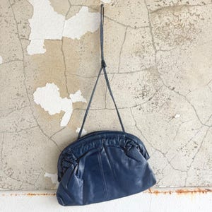 Könnte beinhalten: Eine dunkelblaue Handtasche aus Leder mit einem abgerundeten, gerafften Verschluss und einem dünnen, langen Schulterriemen. Die Tasche hat eine glatte Oberfläche am Hauptteil und ein strukturiertes, gefaltetes Detail entlang der oberen Kante.
