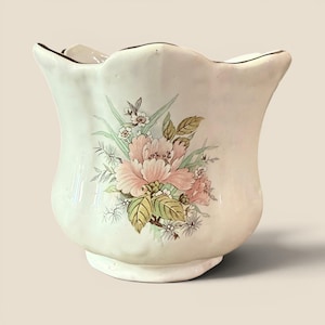 May include: A cream-colored ceramic planter with a scalloped rim and a textured surface. The front features a floral illustration of a large pink peony surrounded by green leaves and smaller white blossoms, with a dark brown trim along the top edge.